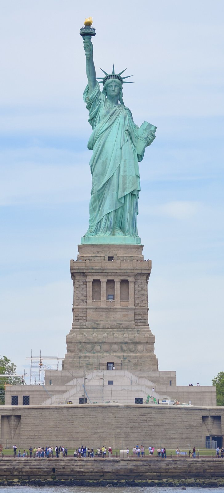 The Statue of Liberty seen from the water, standing on its tall granite pedestal above the star-shaped fort walls, with visitors along the shore